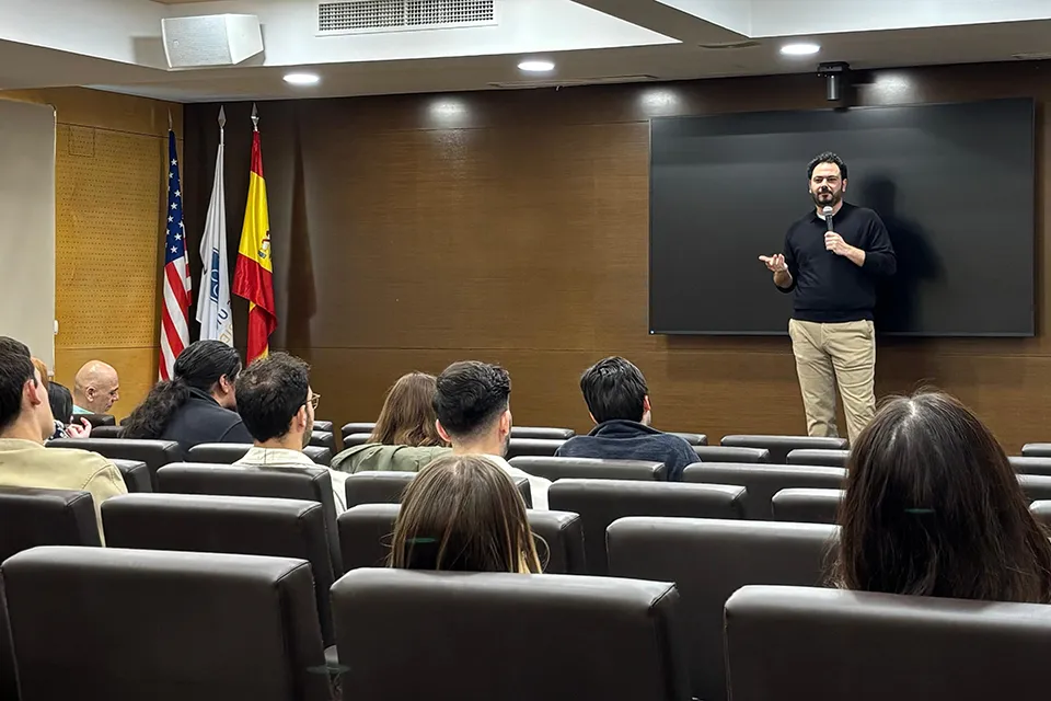 Nehme presenting in the San Ignacio Hall Auditorium on February 25. People sitting in brown auditorium chairs look at the speaker on stage who is speaking into a handheld microphone with the American and Spanish flags on the stage to the left.