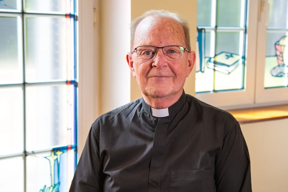James O’Leary, S.J., photographed in the PRH Chapel. Priest poses for photo with stained glass windows in the background with religious designs.