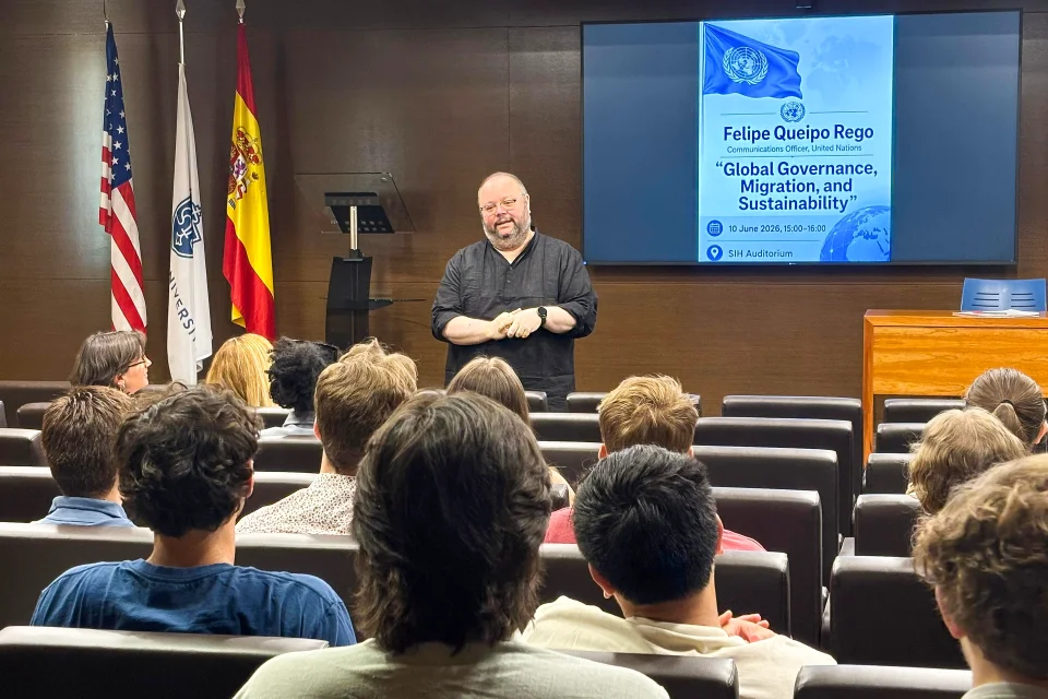 Man speaking to students with flags of the U.S., the University, and Spain to the left and the poster of the event is projected on the screen to his right.