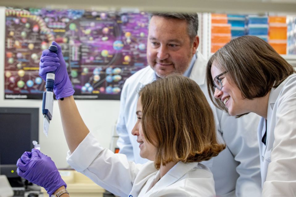 Three medical researchers in white labs collaborate in a lab, with two smiling and watching while the other uses a pipette.