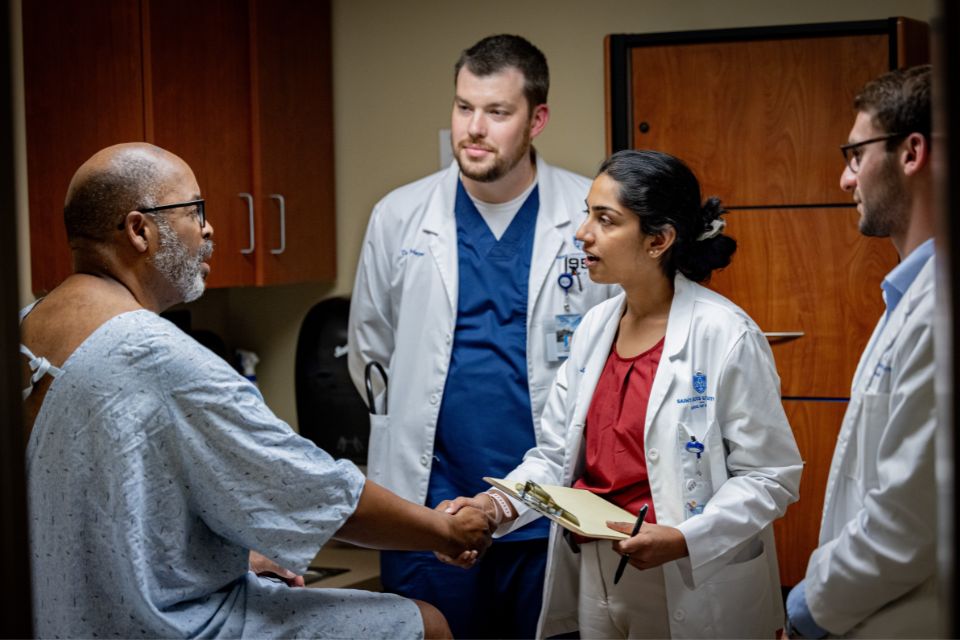Two medical students in white coats interact with a patient in a hospital gown.