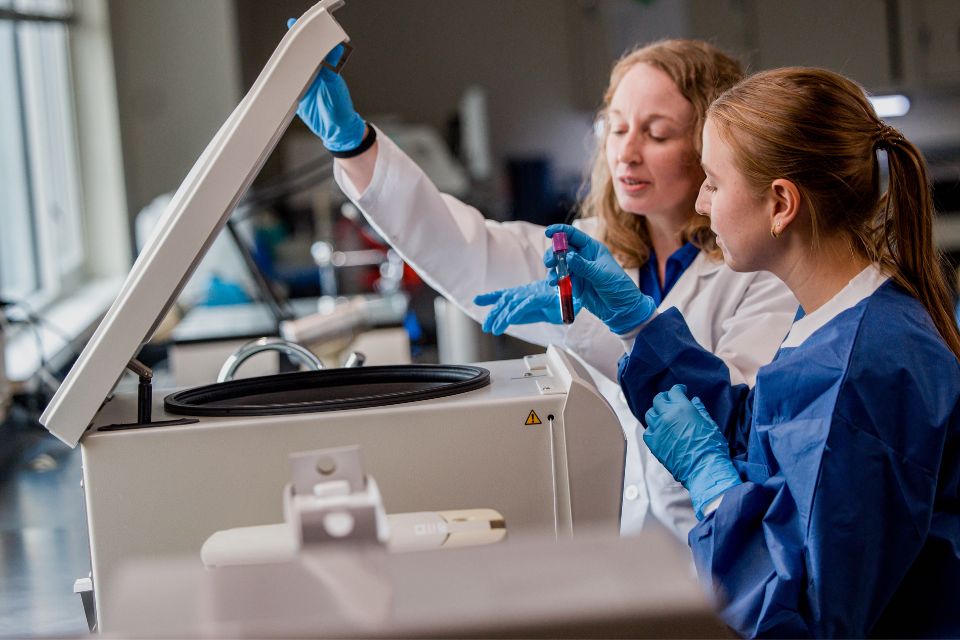 A researcher wearing a lab coat and a student wearing scrubs process a blood sample inside a laboratory.