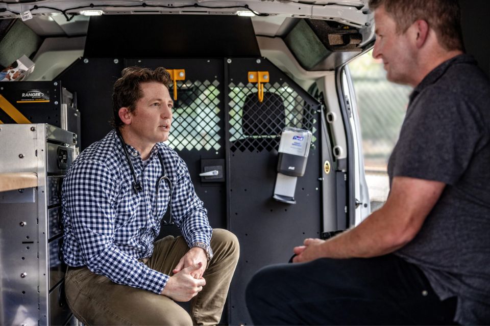 A man wearing a stethoscope speaks to a patient inside a mobile medical van.