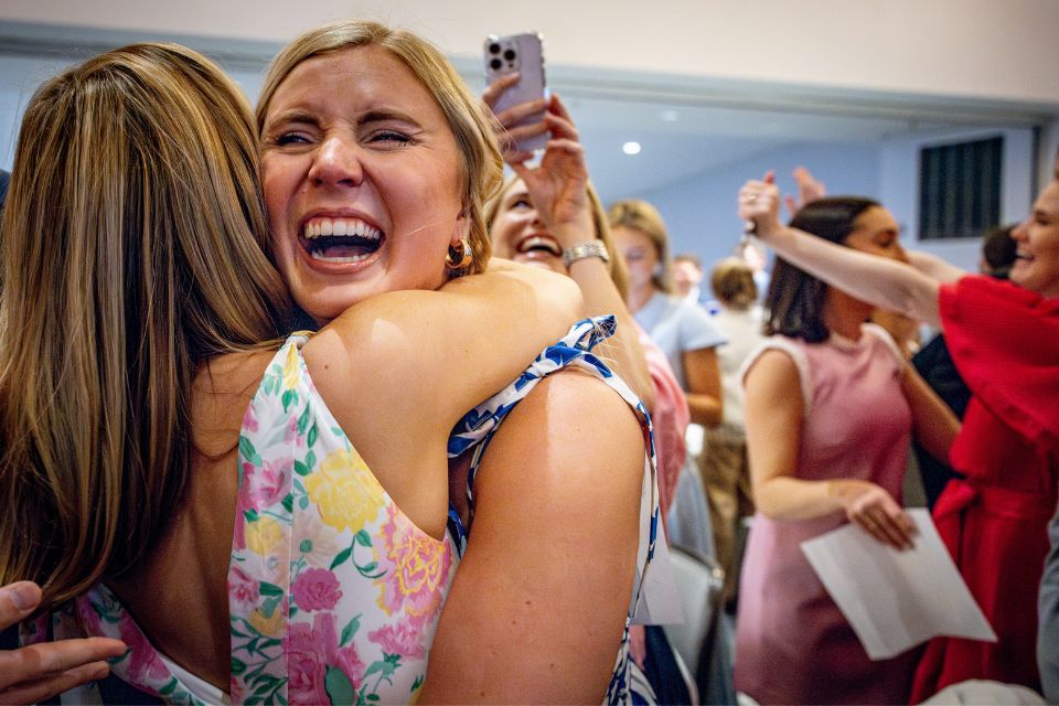 Two smiling women hug one another excitedly in a room filled with people celebrating.