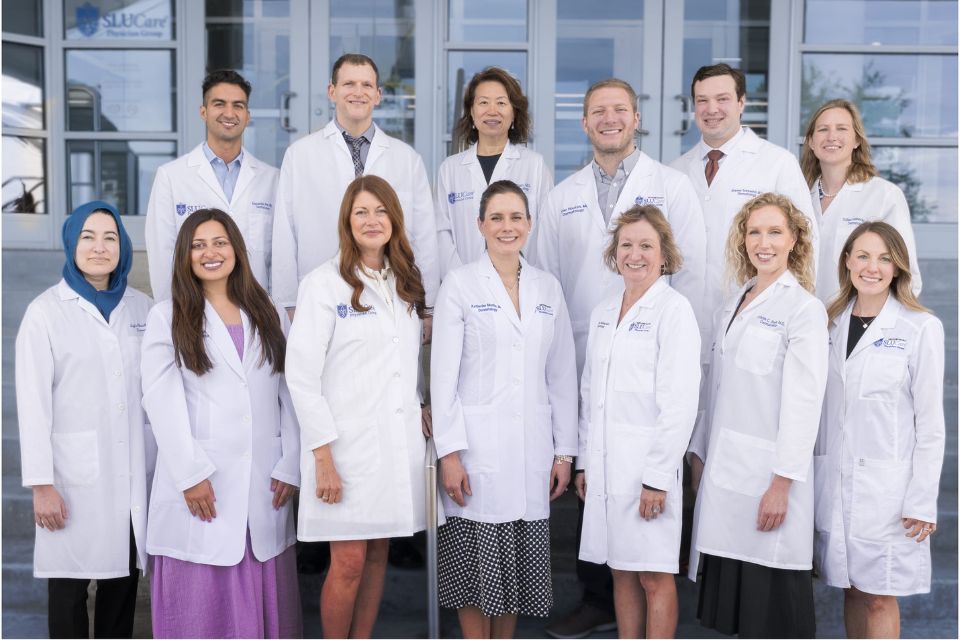 SLU Dermatology Faculty A group of doctors in white coats smile and pose together outside an office building entrance.
