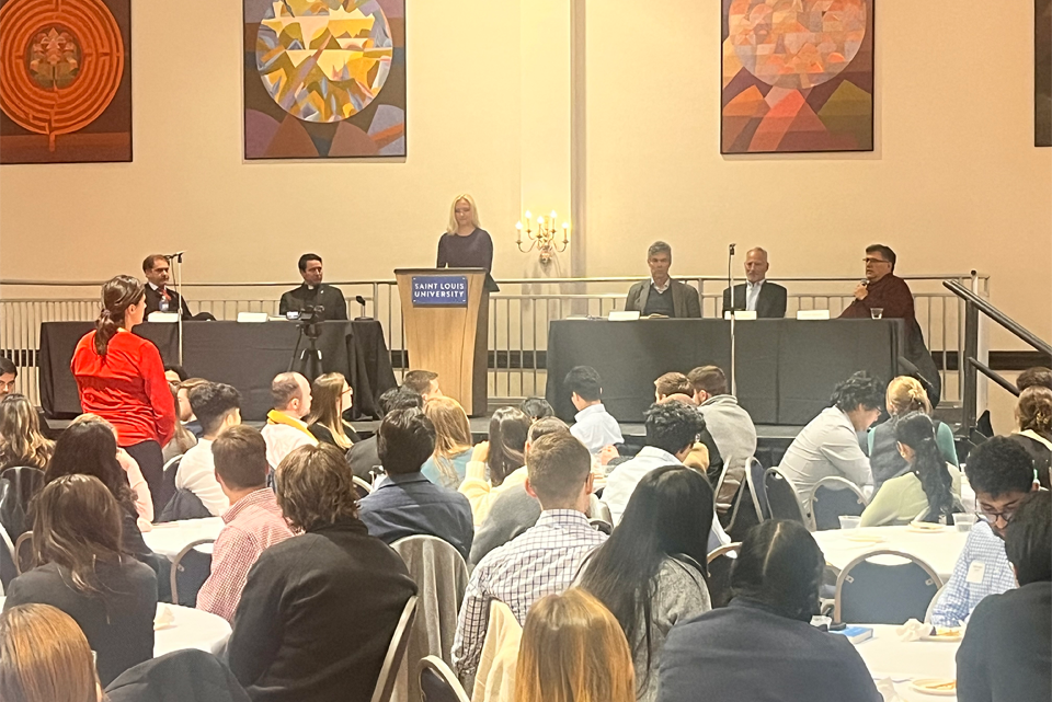 Panelists sit at two tables with a speaker at a lectern in between. The audience, seen from behind, watches while seated at round tables.