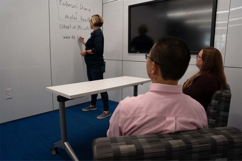 two students being instructed by a professor in front of a whiteboard.