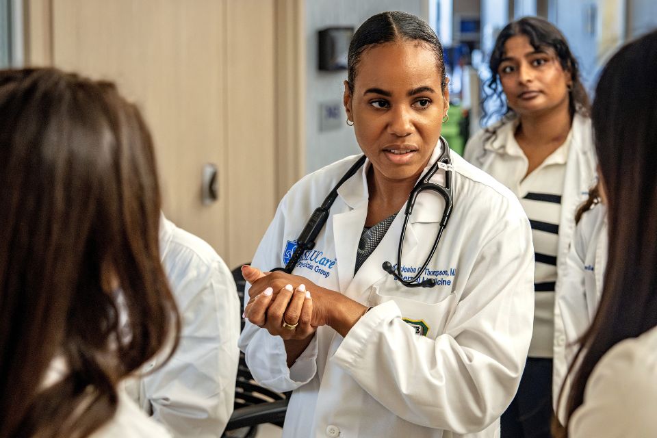 A group of internal medicine residents stand in a hospital hallway listening to a physician. 