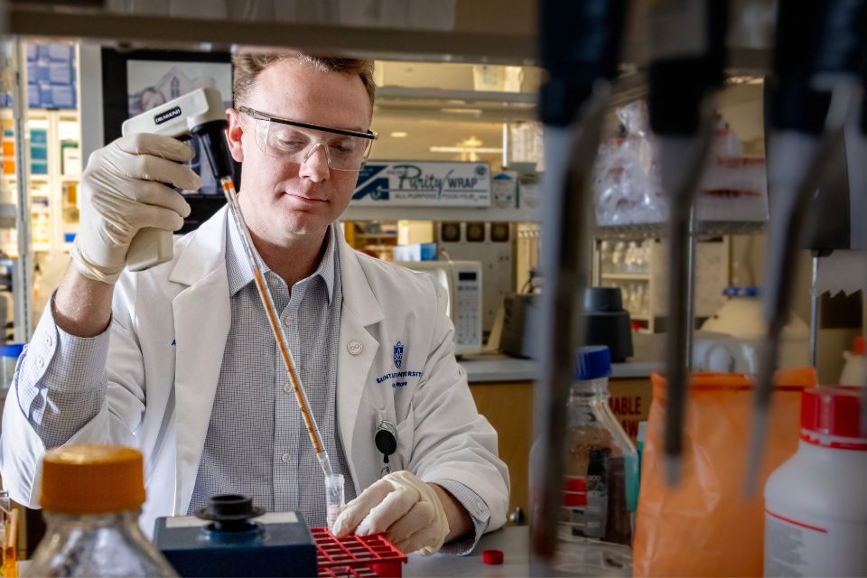A scientist wearing safety glasses and a lab coat uses a pipette in a research lab.