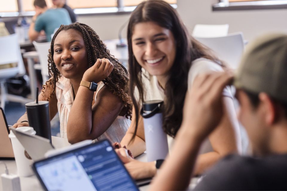 Smiling medical students sit at a classroom table with water bottles and open laptops.