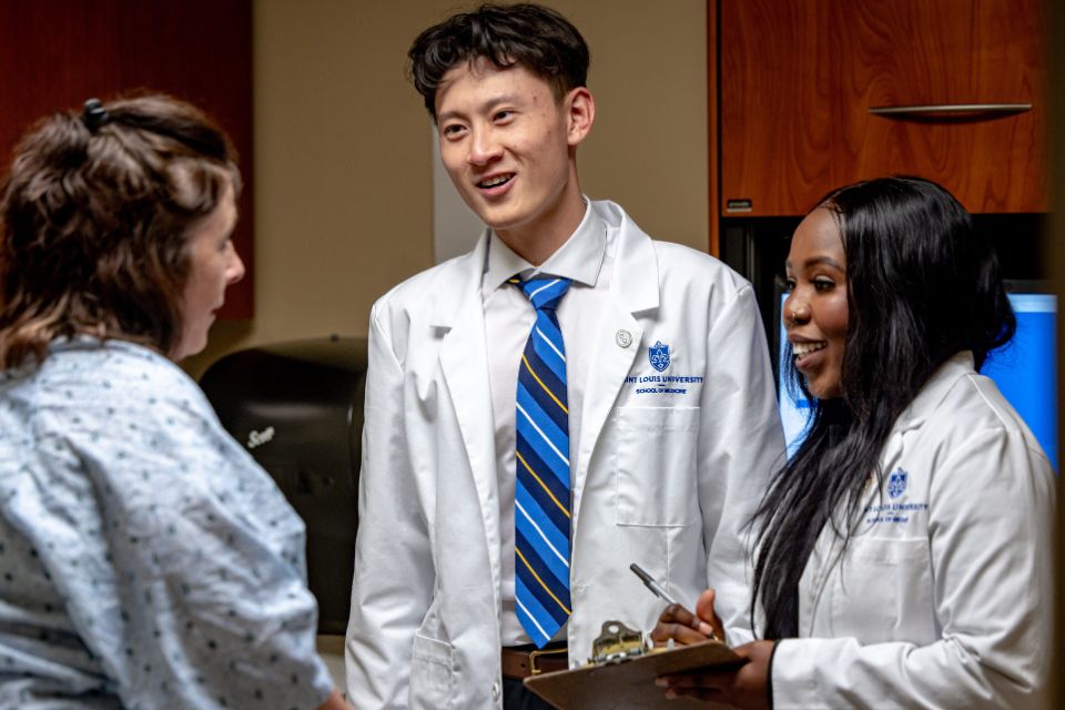 Two medical students in white coats greet a patient sitting in an exam room.