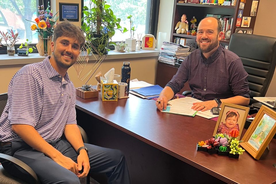 A smiling man sits behind an office desk across from another smiling student.