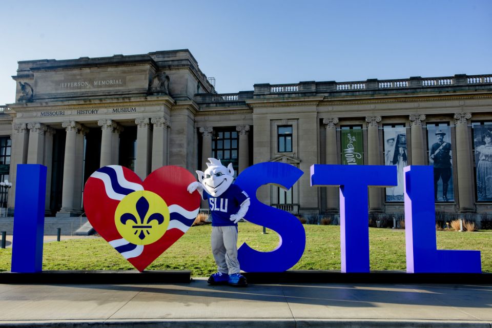 The SLU Billiken poses next to a large "I Love STL" sign outside the Missouri History Museum entrance.