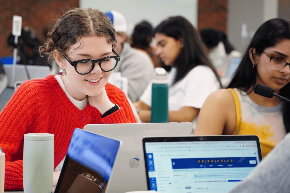 A medical student sits at a desk smiling while looking down at an open laptop screen.