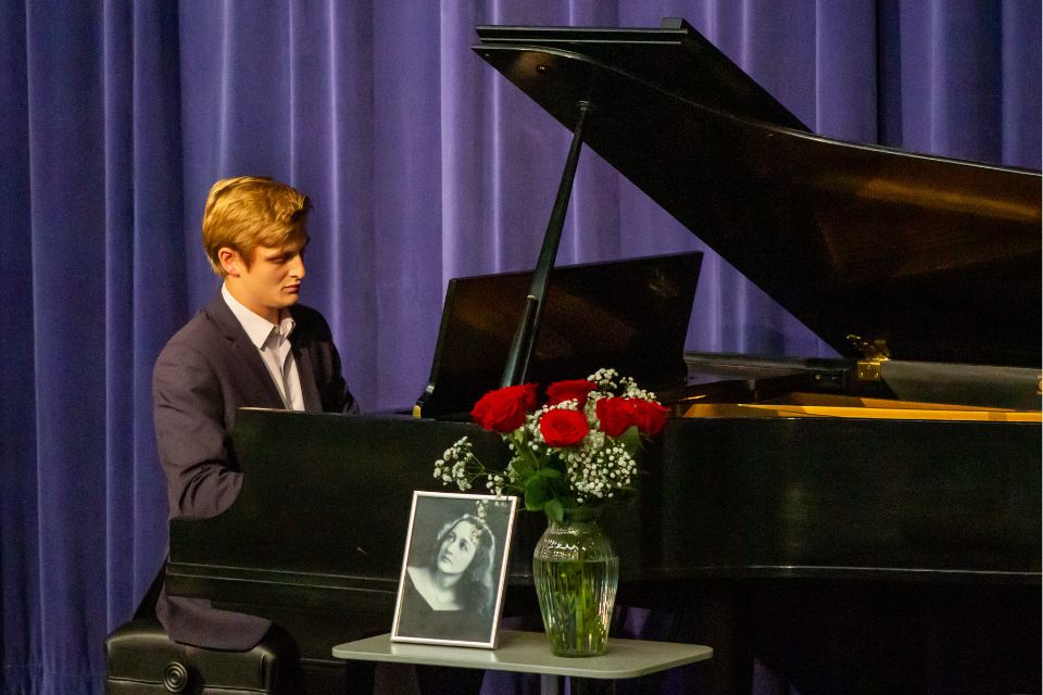 A student in a suit plays a grand piano on stage next to a vase of flowers and a framed black and white portrait of a woman.