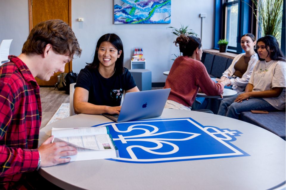 Medical students sit at a table with an open book and laptop screen while another group talks on a nearby couch.