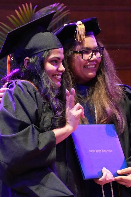 Two people in caps and gowns smile while holding a SLU SOM degree.