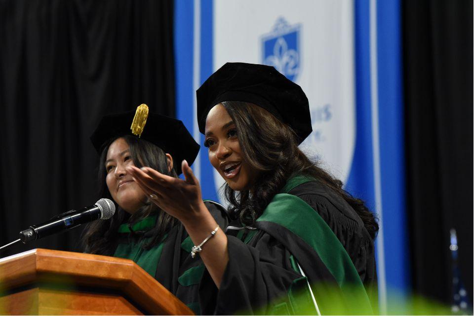 Saint Louis University School of Medicine M.D. Commencement Ceremony Two students dressed in caps and gowns stand at a podium in front of a blue banner and to address an audience.