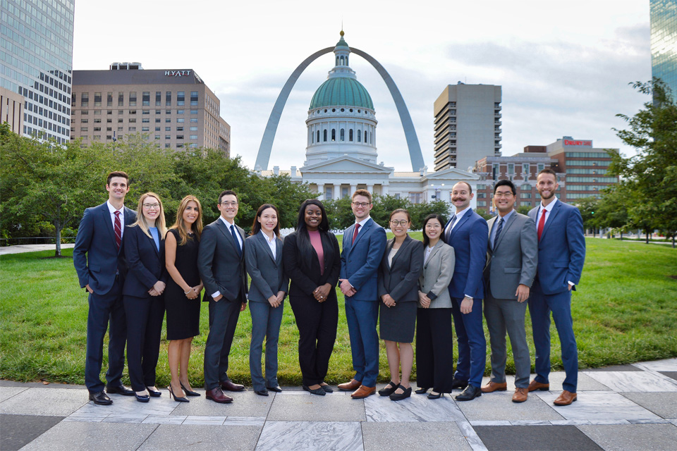 A group of residents wearing suits stand in a row with the St. Louis arch and Old Courthouse in the background.