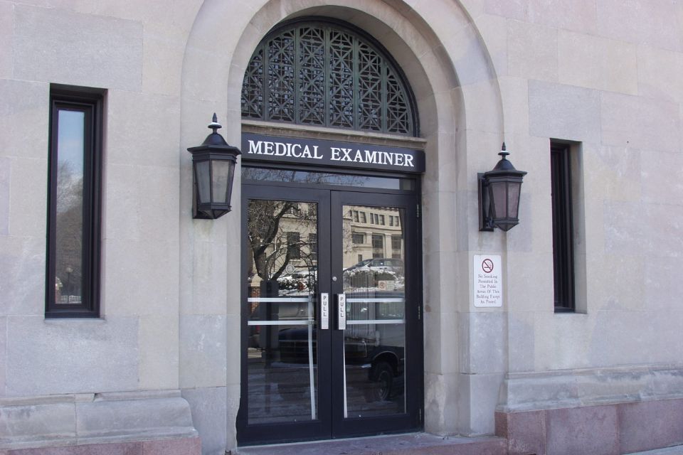 An entrance to the medical examiner office that is stone with double glass doors and an arched window.
