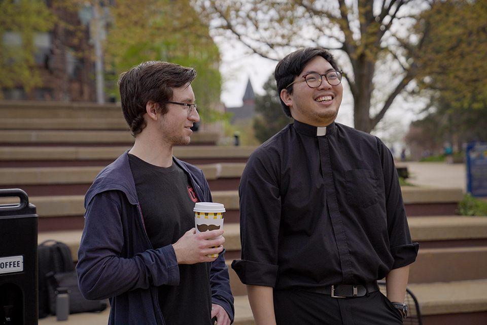 A priest stands next to a man attending Java with the Jesuits. They're talking and smiling.