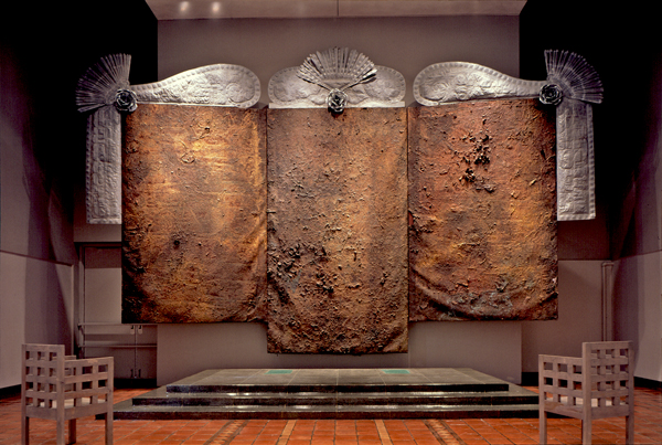 An artwork by artist Michael Tracy titled, Triptych-11th, 12th, and 13th Stations of the Cross for Latin America. Three large vertical panels are joined together like an altarpiece. The central panel is longer and extends lower than the two side panels. All three are painted in earth tones and are highly textured, with prominent creases and tears. Hammered tin panels like fans and curtains are mounted on the top and sides of the work.