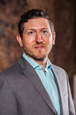 David Brinker MOCRA Director David Brinker, wearing a light blue dress shirt and grey sportcoat, poses in front of an abstract brown background