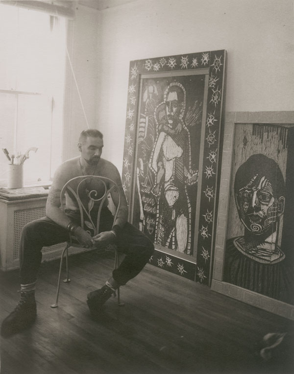 A sepiatone photograph of artist Adrian Kellard in his studio. Kellard is seated in a chair with his back to a sunlit window. Two large woodcut panels lean against the wall nearby.