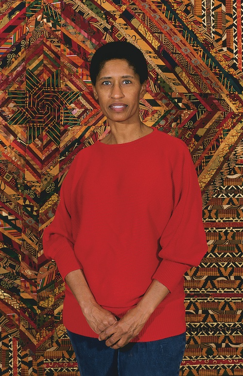 Artist Gwendolyn A. Magee stands in front of an intricately patterened fabric wall hanging