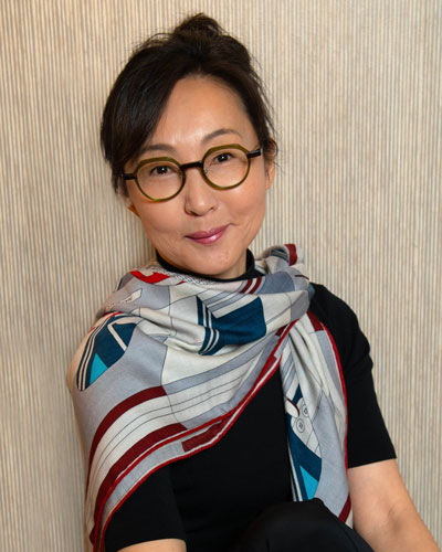 Performance artist Janet Park smiles wryly at the viewer. She wears a black shirt with a scarf bearing geometric patterns in grays, blues, and reds.