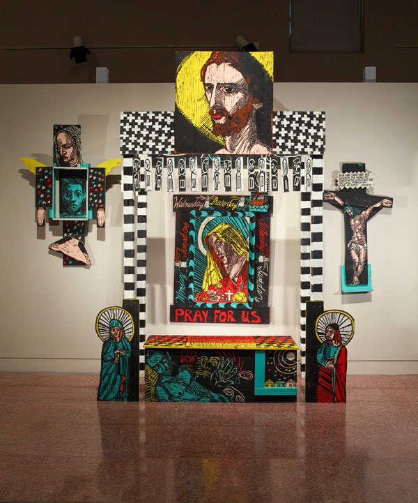 An artwork by Adrian Kellard titled Healing . . . The Learned Art of Compassion. A large wooden canopy stands over a wooden altar. The structure is carved and paninted in yellow, teal, red, black, and white. Various images include the face of Christ, Mary, St. John, a reclining figure of a nude male, and various geometric patterns. Centered on the wall behind is a carved wooden image of Mary with the words, "Pray for Us." To one side hangs a wooden sculpture featuring a self-portrait of the artist embraced by a winged Mary figure, and to the other side a crucifixion. 