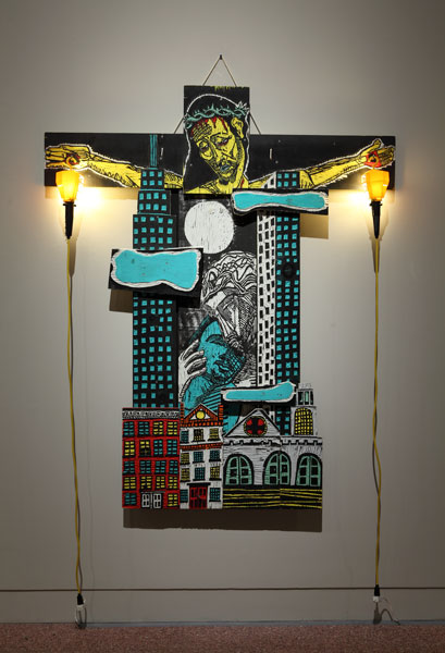 An artwork by Adrian Kellard titled Lovers. A sculptural work made up of various carved wood panels. At top, the head and arms of Jesus on the Cross. In the place of his body, a cityscape with skyscrapers and, at bottom, a street scen with apartment buildings and a church. In the middle of the work, between the skyscrapers, a man embraces another man tenderly. Yellow trouble lamps are suspended from the hands of Christ, with the power cords dangling down.