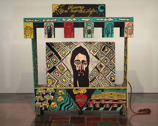 An artwork by Adrian Kellard titled Shrine. A large wagon made of carved wood panels painted in yellow, teal, red, and black, features carved drawings including the face of Jesus, the Sacred Heart, buildings and a landscape, whimsical angels, and the words, "Rising You restored Life."