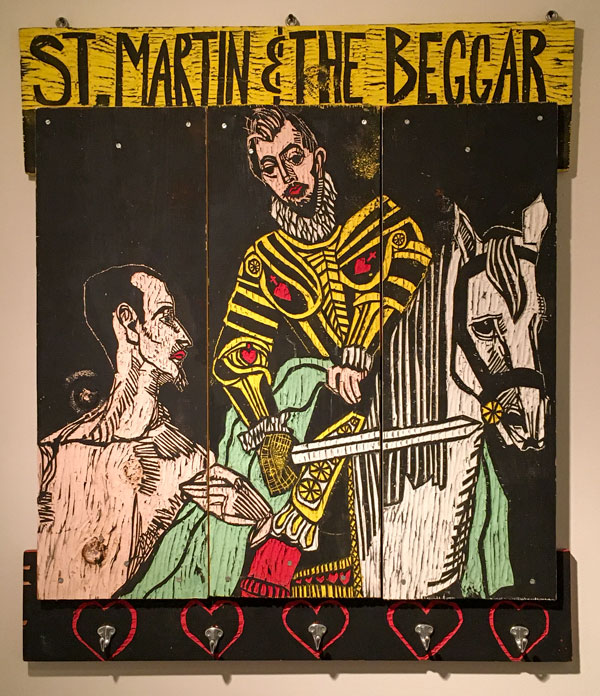 An artwork by Adrian Kellard titled St. Martin of Tours coat rack. A large carved wood panel features a visual quotation of a painting by El Greco in which St. Martin, seated on a white horse, cuts his cloak in half to get part to a naked beggar standing below to the left. At top are the words, "St. Martin & the Beggar." Along the bottom, mounted inside carved hearts, are hanging hooks.