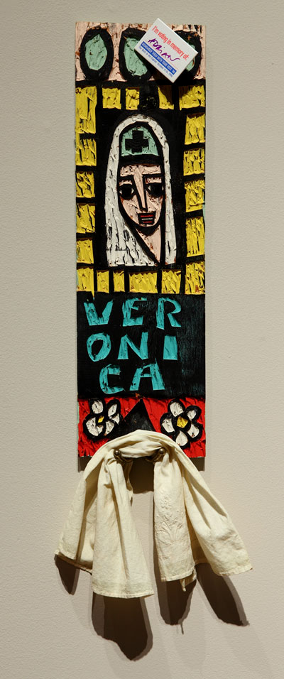 An artwork by Adrian Kellard titled Veronica towel rack. A narrow rectangular carved piece of wood features the face of Veronica with her name below, various geometric patterns, and a hook with a towel hanging from it. The work is painted in black, yellow, teal, red and white. 