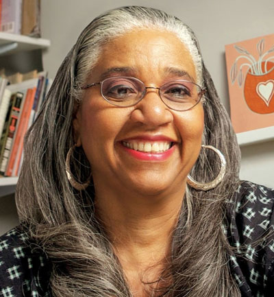 Curator and art historian Leslie King-Hammond, wearing large silver hoop earrings, sits in an office with books and a small painting in the background