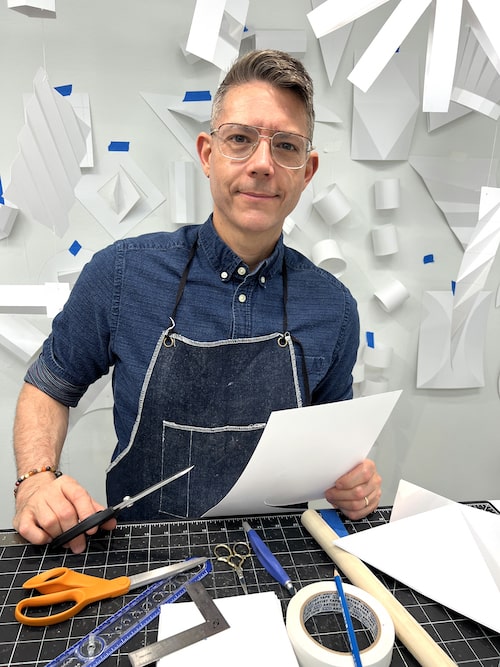 Artist Michael Velliquette, wearing a denim smock, stands in his studio surrounded by his tools and work in progress