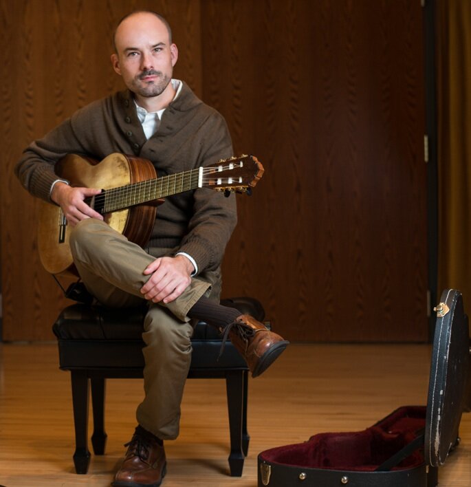 Guitarist Patrick Rafferty sits on a piano bench with his guitar on his lap, an open guitar case on the floor next to him