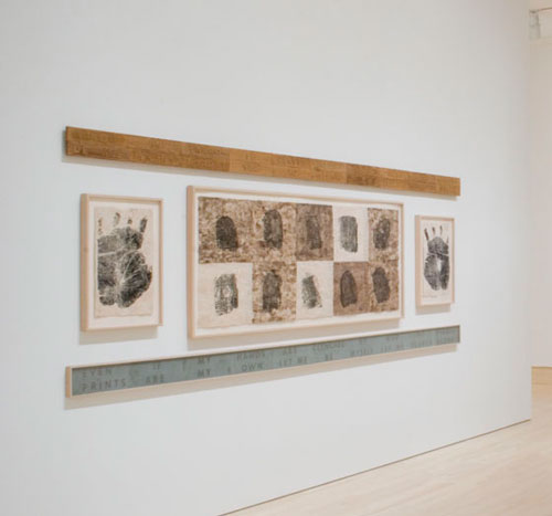 An artwork by Maria Magdalena Campos-Pons titled, Birth Certificate. Multiple photographs of handprints and fingerprints are arranged on a wall.