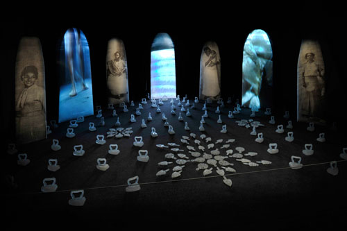 An installation by Maria Magdalena Campos-Pons titled, Spoken Softly with Mama. Ironing boards are leaned upright against a wall in a semi-circle. Photos of women and landscapes are projected on them. Arranged in front of them on the floor are old-fashioned flat irons and trivets.
