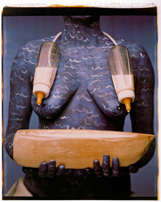 An artwork by Maria Magdalena Campos-Pons titled, When I Am Not Here / Estoy Allá. A photograph of the torse of a woman who is painted in blue with white waves, holding a stylized wooden boat. Two baby bottles half-filled with milk are connected by a cord behind her neck and suspended over her chest.