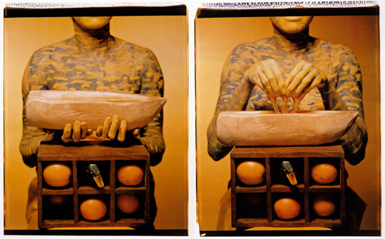 An artwork by Maria Magdalena Campos-Pons titled, When I Am Not Here / Estoy Allá. Two photographs of the torso of a women, her skin painted blue with white waves, kneeling behind a box with six cubbies containing various objects. In the left panel, she holds a stylized wooden boat. In the right panel, honey drips into the boat from her fingers.