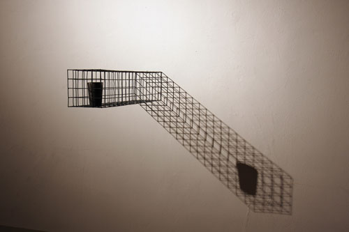 A work by artist Thomas Skomski titled Promise. A cylindrical cuplike object rests at the end of a rectangular cage that extends from a wall. The cage and cup cast a shadow diagonally down to the right.