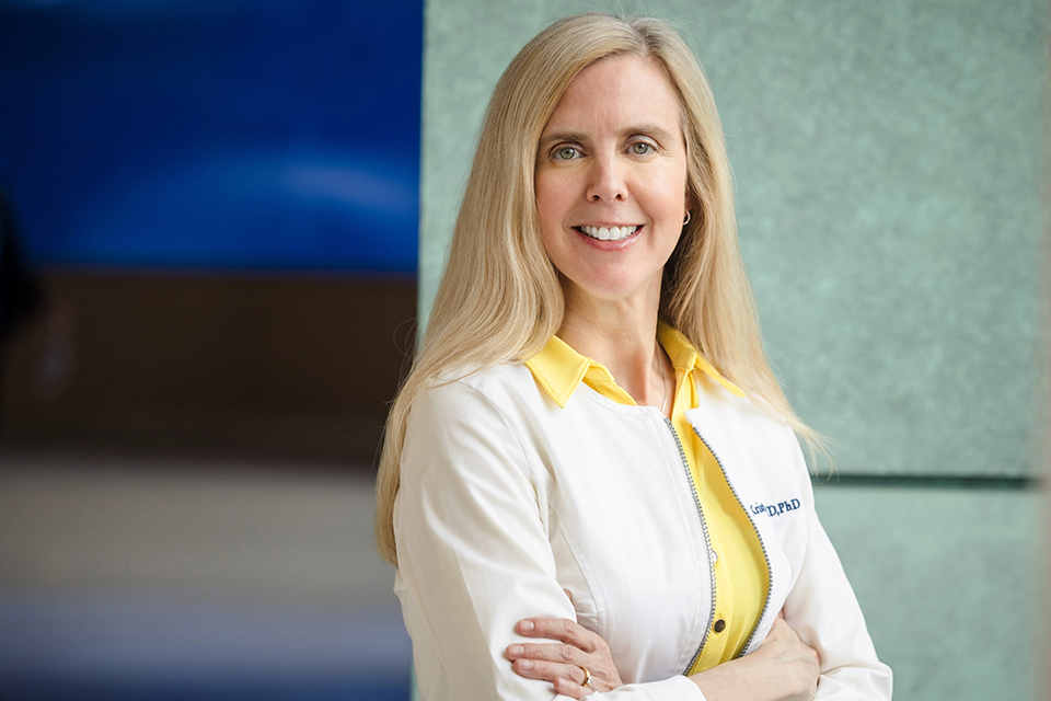 A photo of Dr. Krista Lentine shot indoors. She wears a white lab coat. Her arms are folded across her chest. She smiles at the camera. 