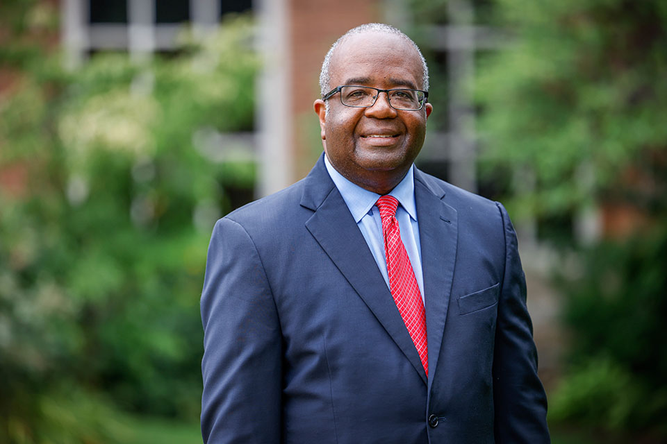 Dr. Travis Threats poses for a photo outdoors during the day. He smiles at the camera. He wears glasses, a blue blazer, a light blue shirt and a red tie. The background is blurred.