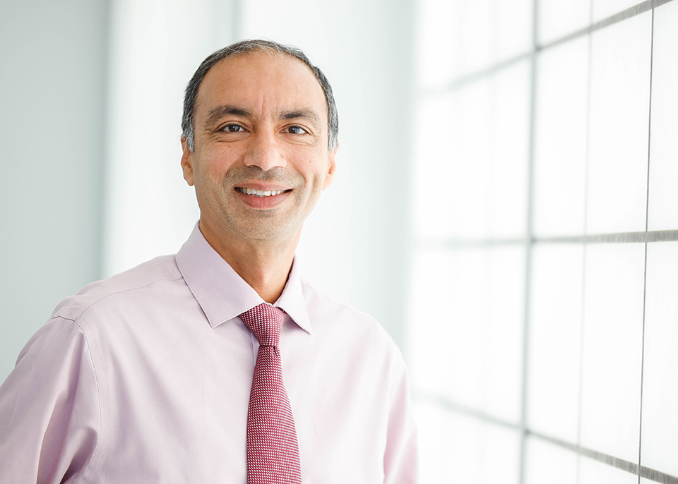Sandeep Dhindsa, M.D. poses for a photo indoors by a window. He wears a pastel pink button-down shirt with a read patterned tie. He smiles for the camera. 