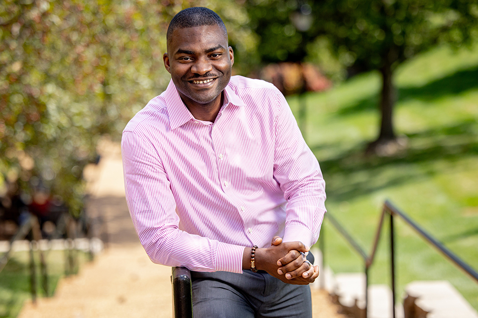 Dr. Oluwatoyosi Owoeye poses for a photo outdoors on SLU's campus on a bright, sunny day. He wears a pink button-down shirt. He smiles at the camera.