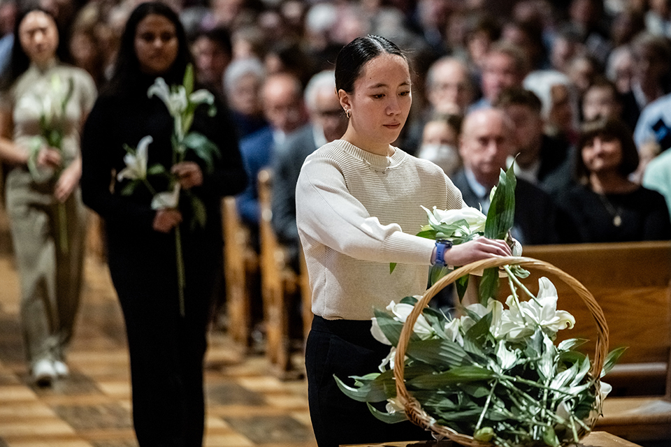A student places flowers into a basket while participants watch from church pews. Students process down the aisle behind her, also carrying flowers.
