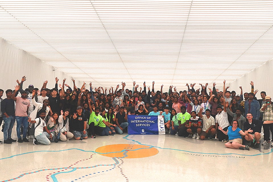 A large group of new international students at SLU raise their hands while posing for a photo in front of the map of the Louisana Purchase on the floor of the Museum at the Gateway Arch. 