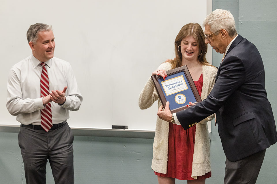 President Fred Pestello hands a plaque to Gabby Chiodo at the front of a classroom while Provost Mike Lewis stands nearby, clapping and smiling.